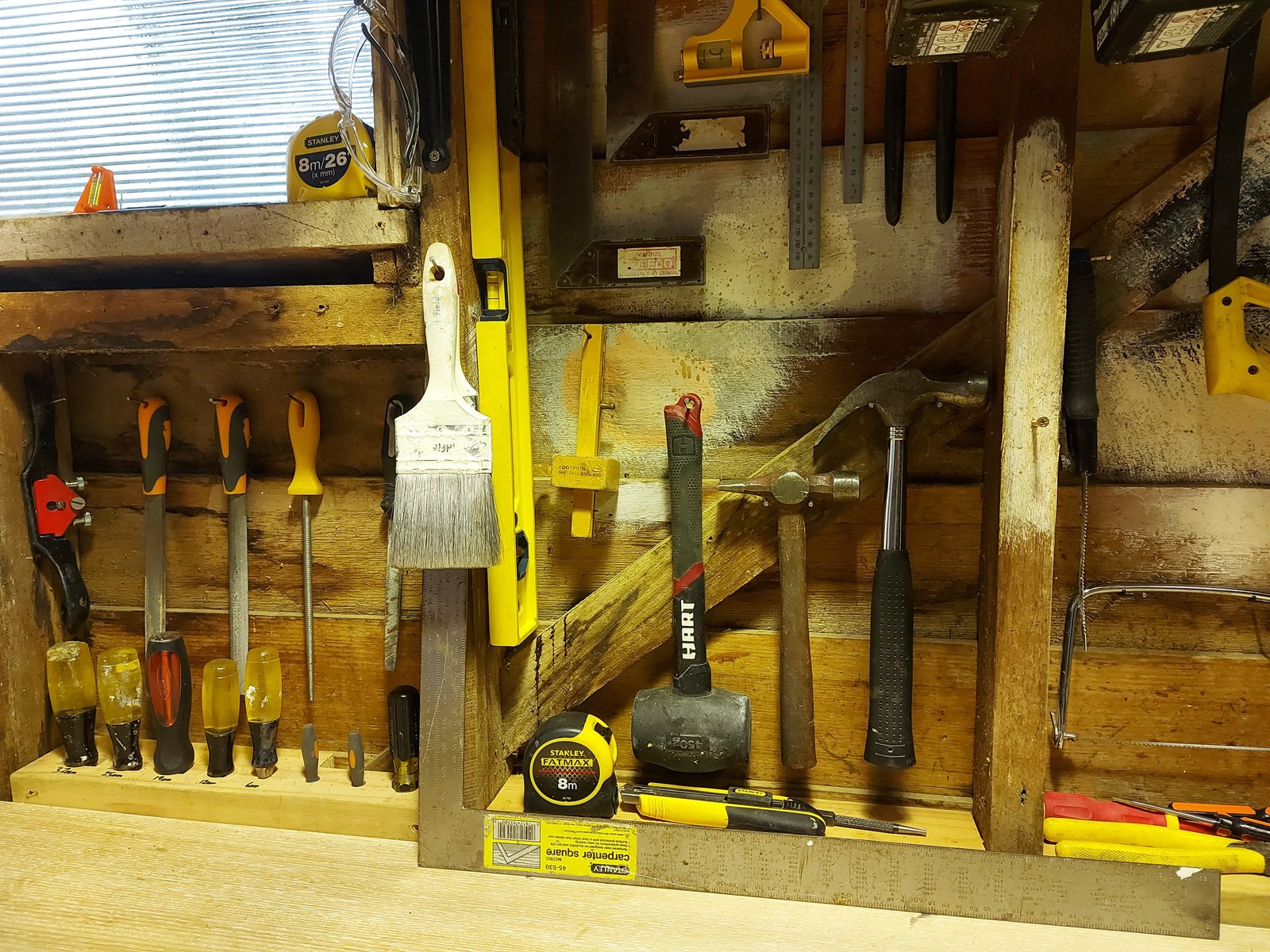 Hand tools including screwdrivers, mallet, hammers, paintbrush, saw, tape measure, and safety glasses on wooden wall.
